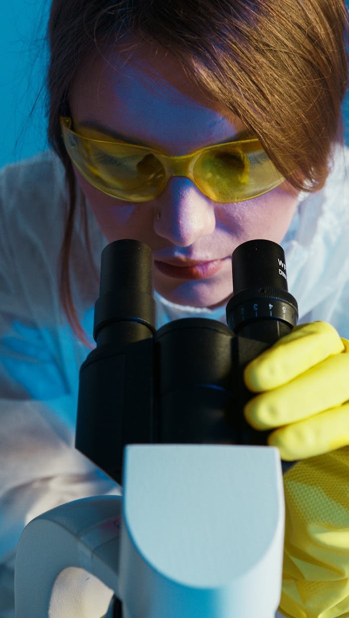 journey A female scientist in a lab coat looks through a microscope wearing yellow protective goggles and gloves.